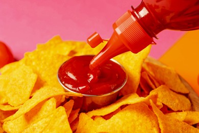 Photo of Pouring tasty ketchup in bowl with nachos on pink background, closeup