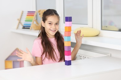 Happy girl with tower of colorful cubes at table indoors