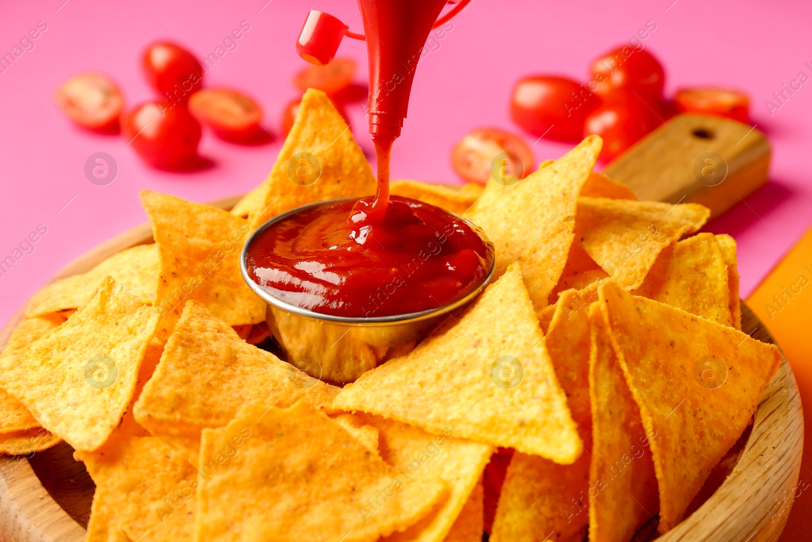 Photo of Pouring tasty ketchup in bowl with nachos on pink background, closeup