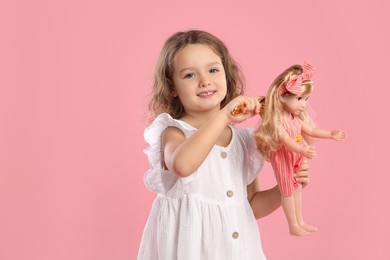 Photo of Cute little girl brushing doll's hair on pink background