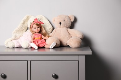 Beautiful doll, teddy bear and toy bunny on grey chest of drawers near white wall