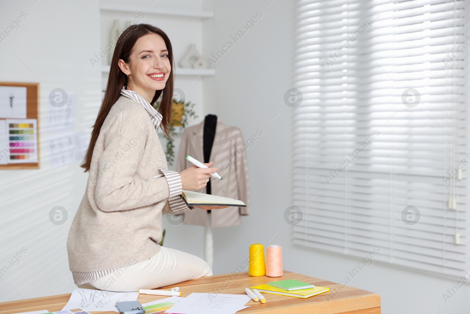 Photo of Fashion designer with marker and notebook sitting on wooden table in workshop, space for text