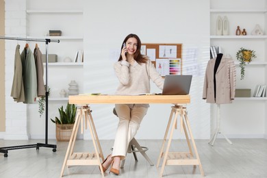 Fashion designer talking on smartphone at wooden table in workshop