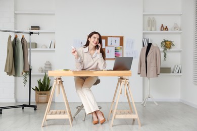 Fashion designer talking on smartphone at wooden table in workshop