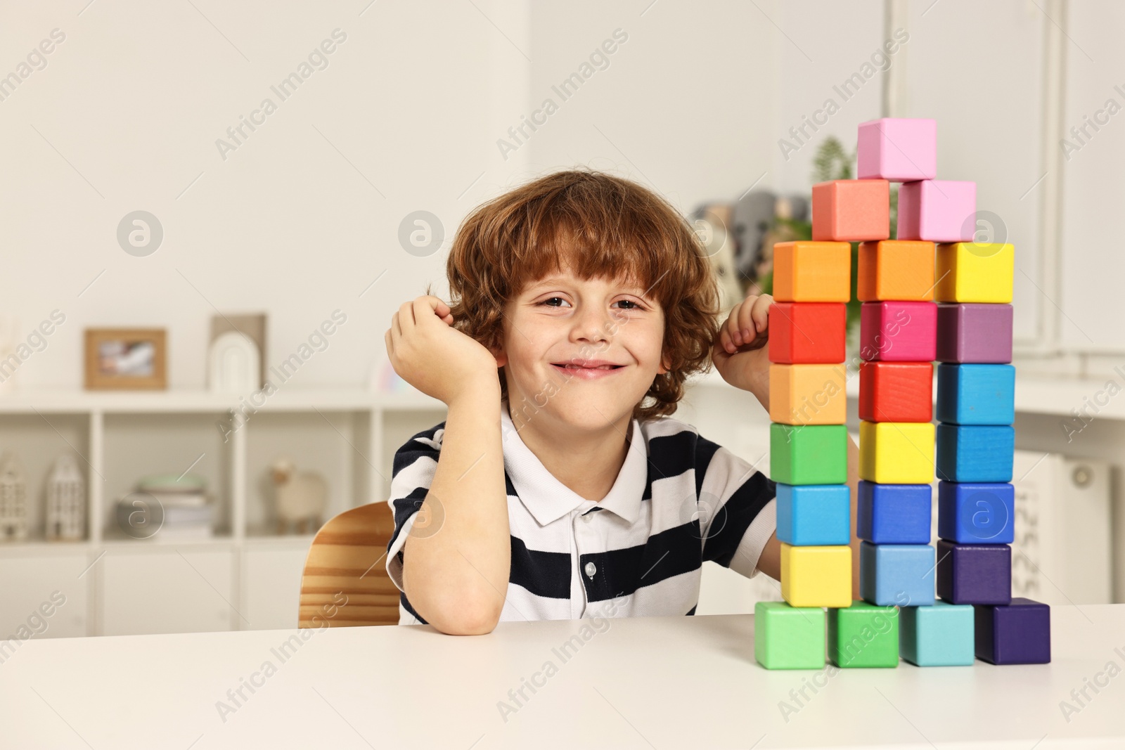 Photo of Little boy playing with stacked colorful cubes at white table indoors