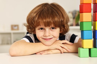 Photo of Little boy playing with stacked colorful cubes at white table indoors