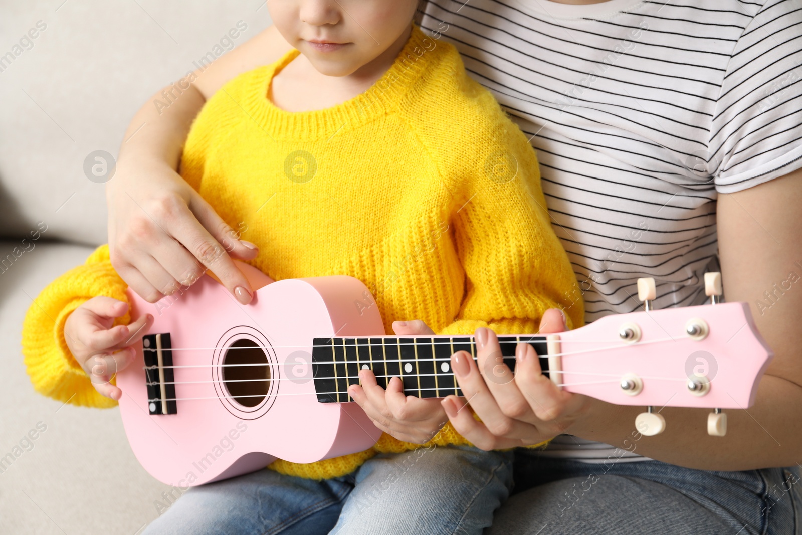 Photo of Woman teaching little girl to play ukulele at home, closeup