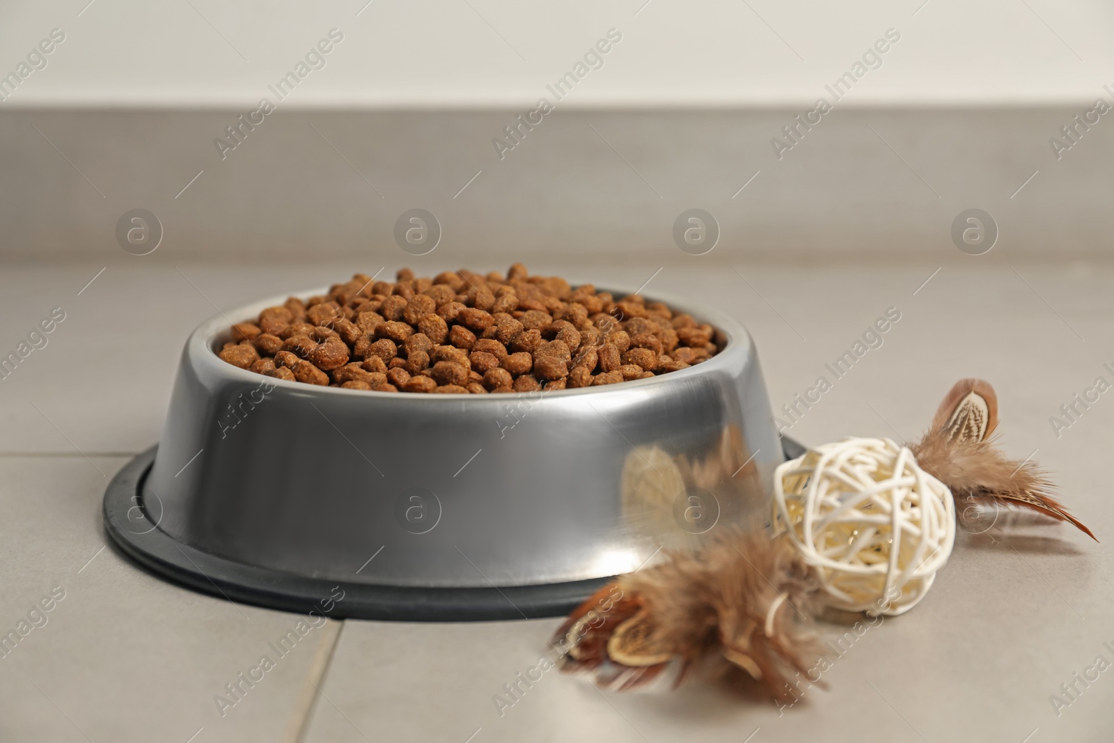 Photo of Dry pet food granules in feeding bowl and cat toy on floor indoors