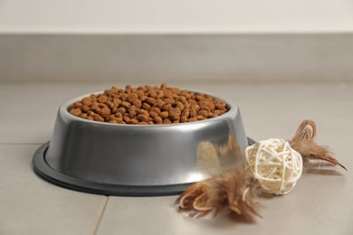 Photo of Dry pet food granules in feeding bowl and cat toy on floor indoors