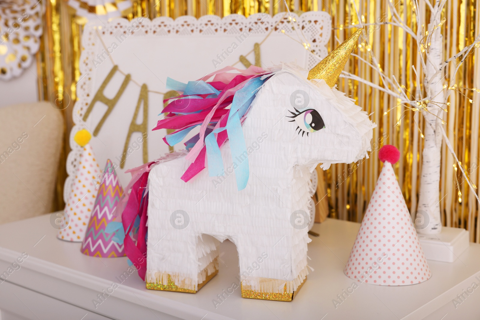 Photo of Bright pinata in shape of unicorn and party accessories on white chest of drawers against foil fringe curtain indoors