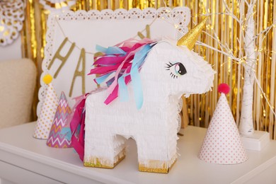 Photo of Bright pinata in shape of unicorn and party accessories on white chest of drawers against foil fringe curtain indoors