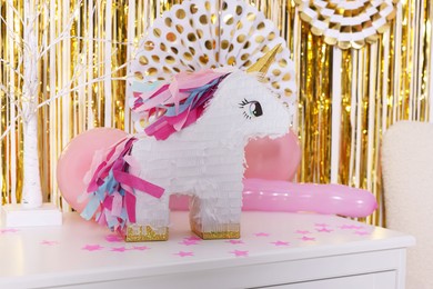 Photo of Bright pinata in shape of unicorn and party accessories on white chest of drawers against foil fringe curtain indoors