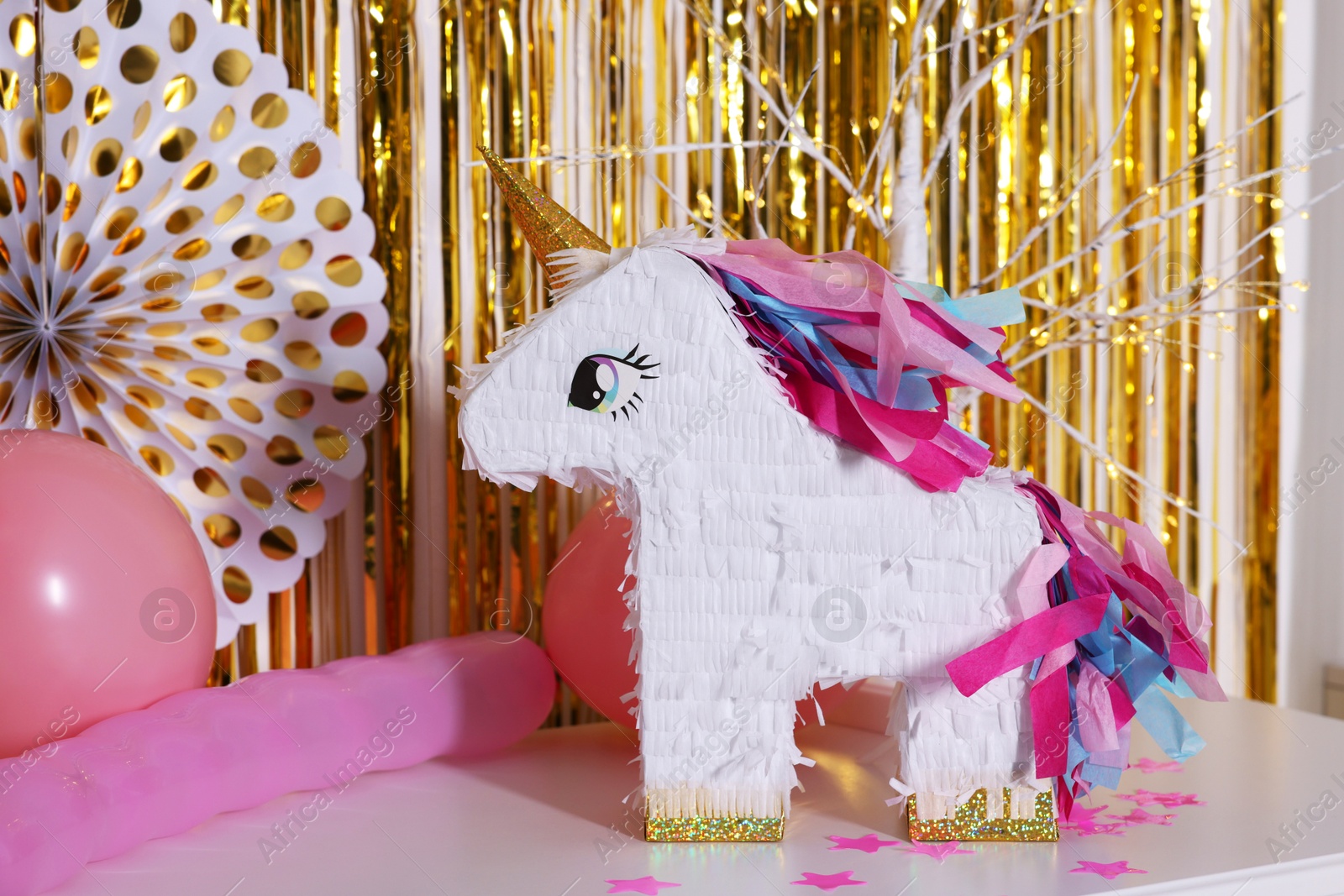 Photo of Bright pinata in shape of unicorn and party accessories on white chest of drawers against foil fringe curtain indoors