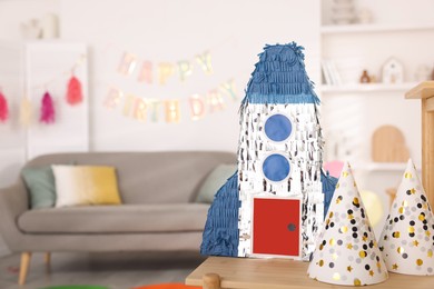 Photo of Bright pinata in shape of rocket and party cones on wooden table in festive decorated room, space for text