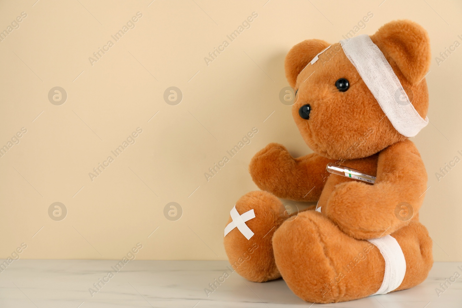 Photo of Teddy bear with bandages, sticking plasters and thermometer on white marble table against beige background, space for text