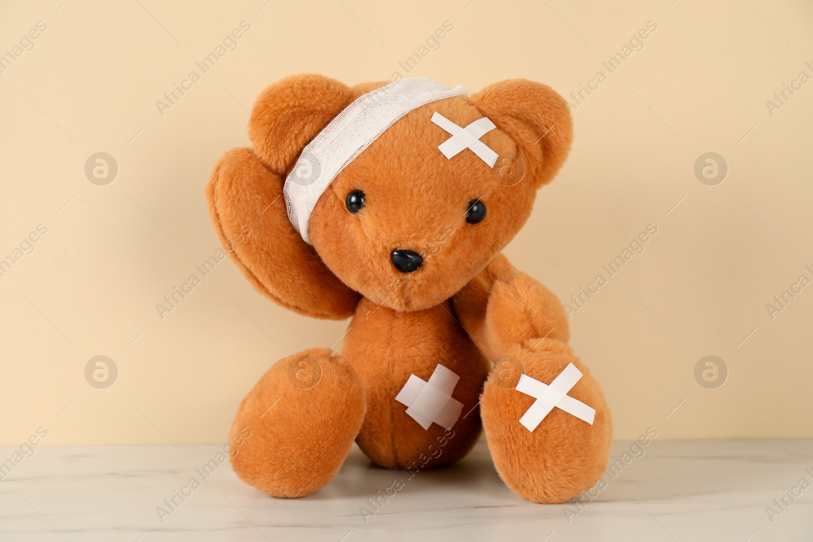Photo of Teddy bear with bandage and sticking plasters on white marble table against beige background