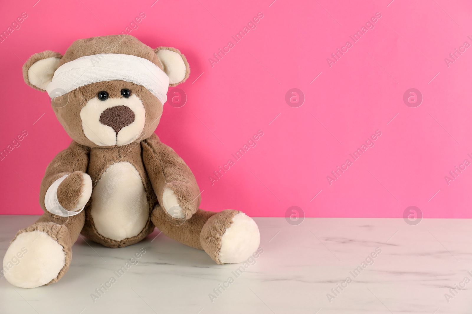Teddy bear with bandages on white marble table against pink background, space for text Photo of Teddy bear with bandages on white marble table against pink background, space for text