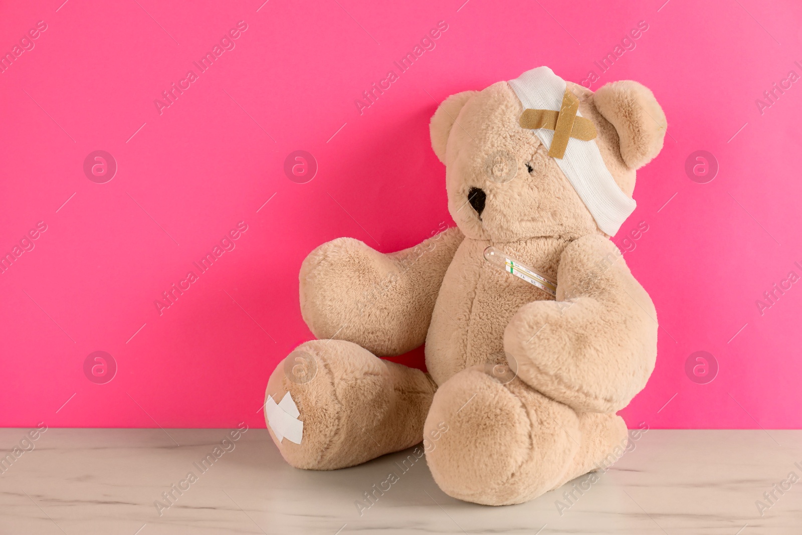 Photo of Teddy bear with bandage, sticking plasters and thermometer on white marble table against pink background