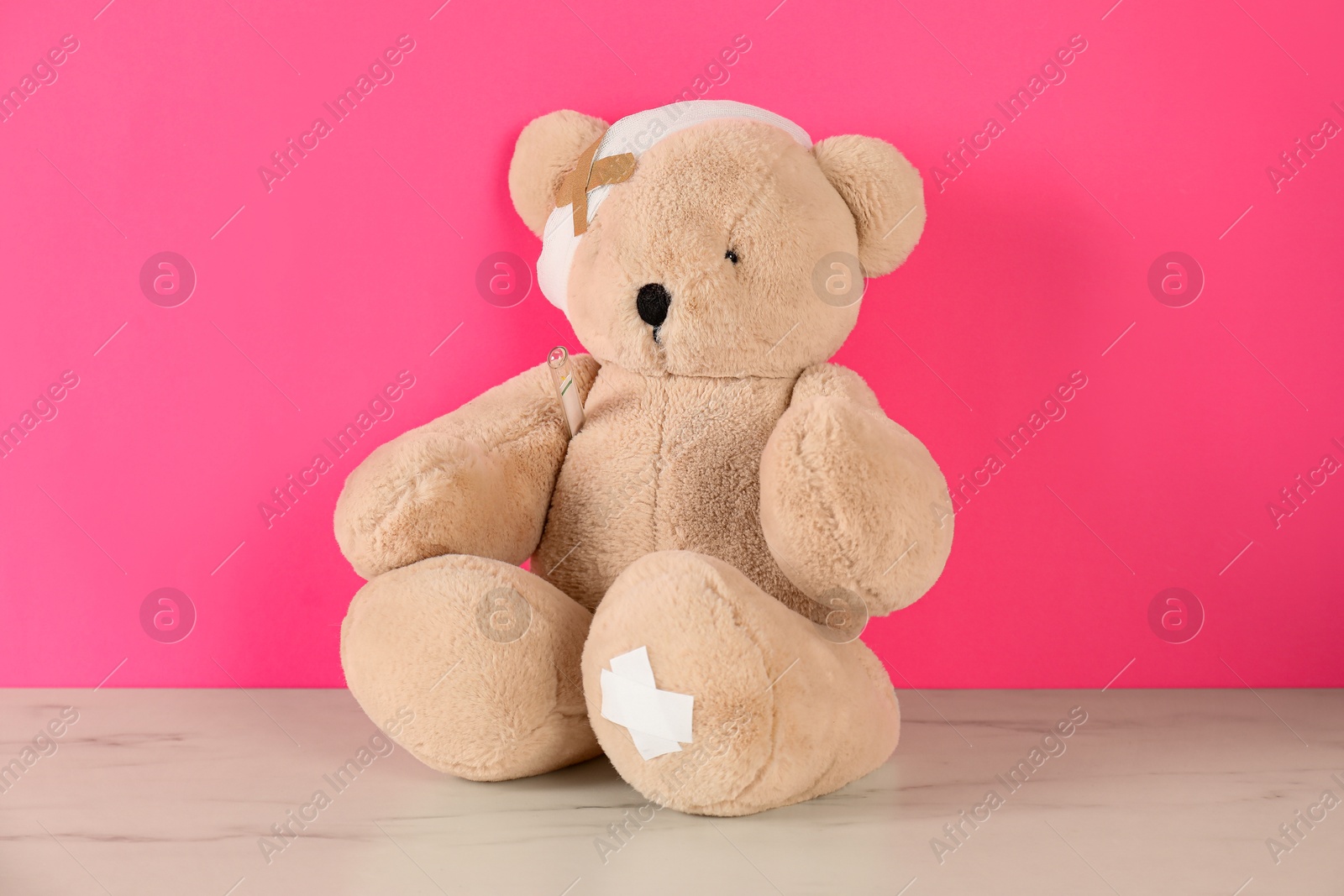Photo of Teddy bear with bandage, sticking plasters and thermometer on white marble table against pink background