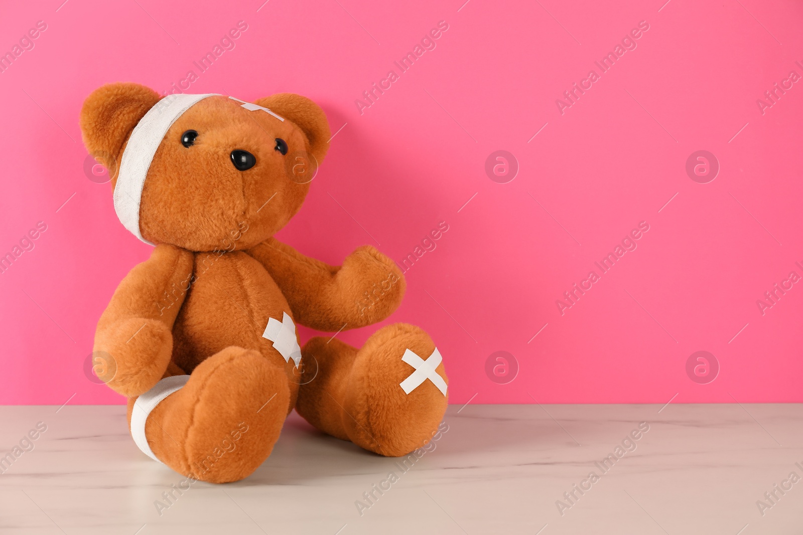 Teddy bear with bandages and sticking plasters on white marble table against pink background, space for text Photo of Teddy bear with bandages and sticking plasters on white marble table against pink background, space for text
