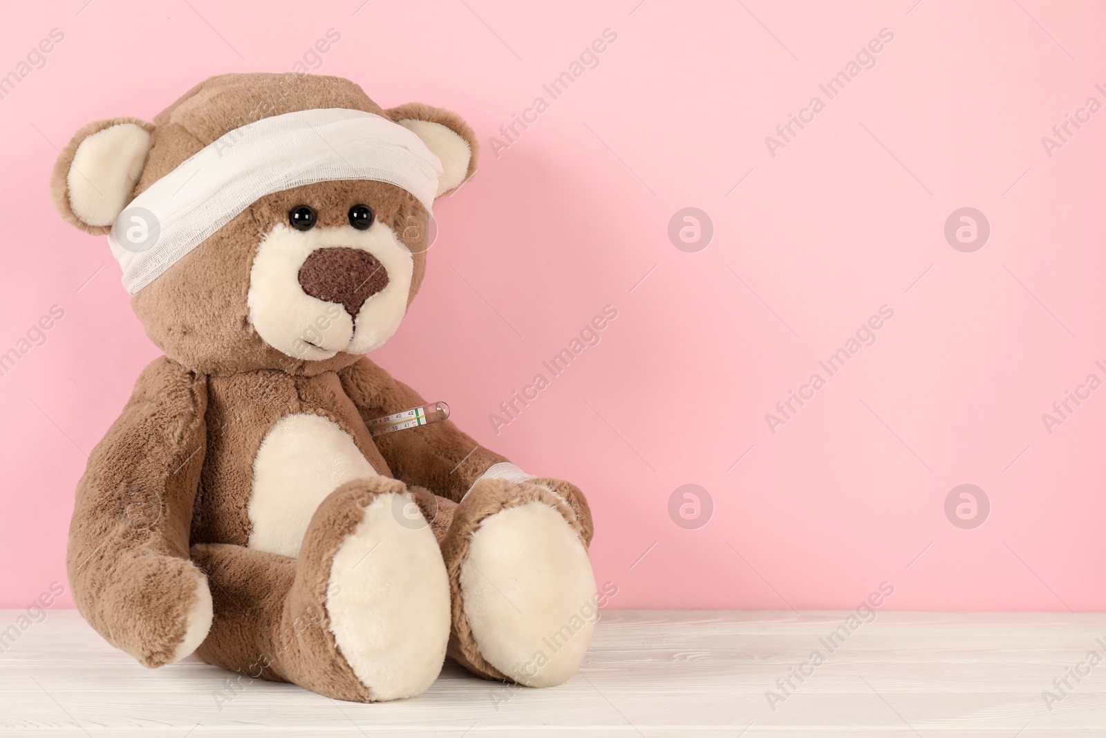 Photo of Teddy bear with bandages and thermometer on white wooden table against pink background, space for text