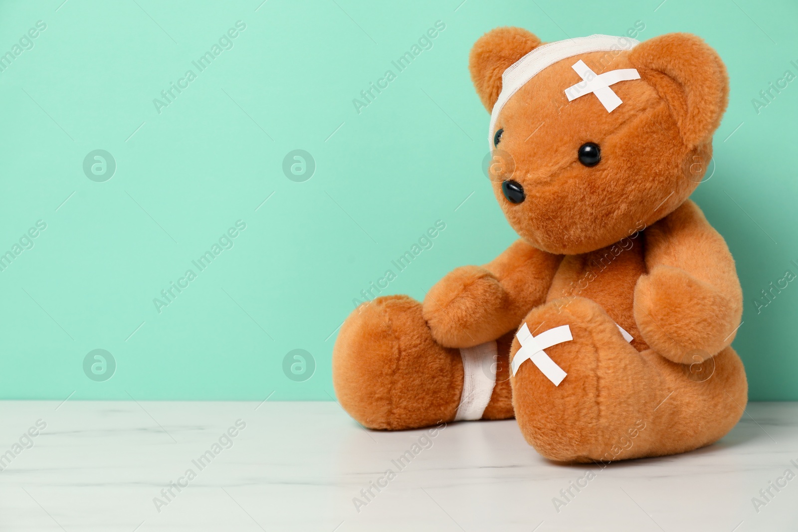 Photo of Teddy bear with bandages and sticking plasters on white marble table against turquoise background, space for text