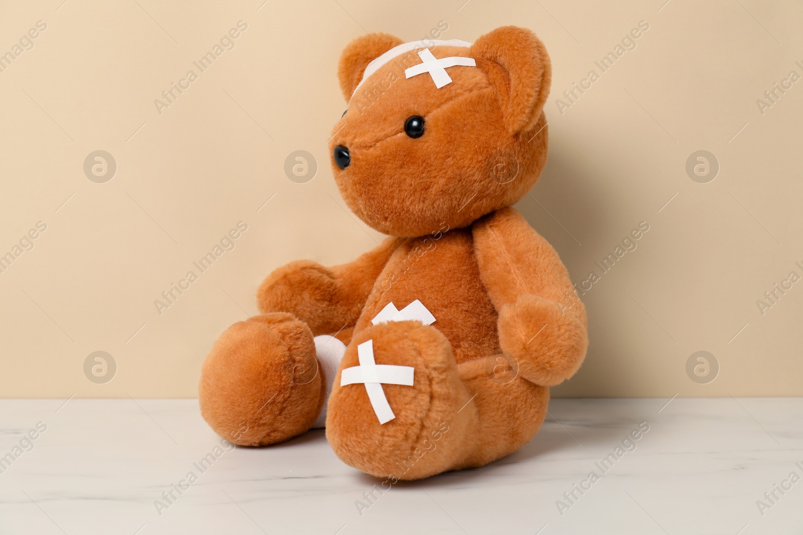 Photo of Teddy bear with bandages and sticking plasters on white marble table against beige background