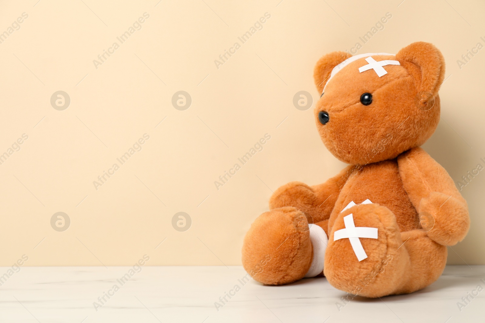 Teddy bear with bandages and sticking plasters on white marble table against beige background, space for text Photo of Teddy bear with bandages and sticking plasters on white marble table against beige background, space for text