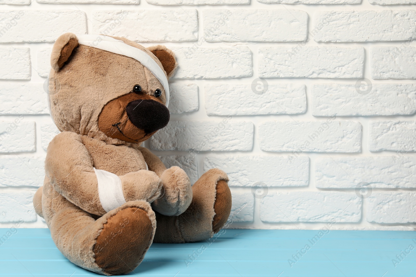 Photo of Teddy bear with bandages on light blue wooden table against white brick wall, space for text
