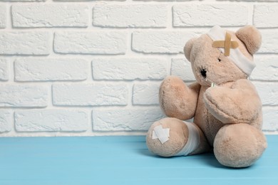 Photo of Teddy bear with bandages, thermometer and sticking plasters on light blue wooden table against white brick wall, space for text