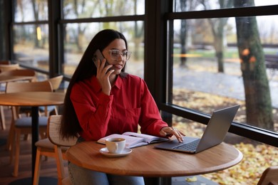 Photo of Woman talking on smartphone while working with laptop at table in cafe