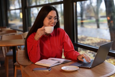 Photo of Woman with cup of coffee working on laptop at table in cafe