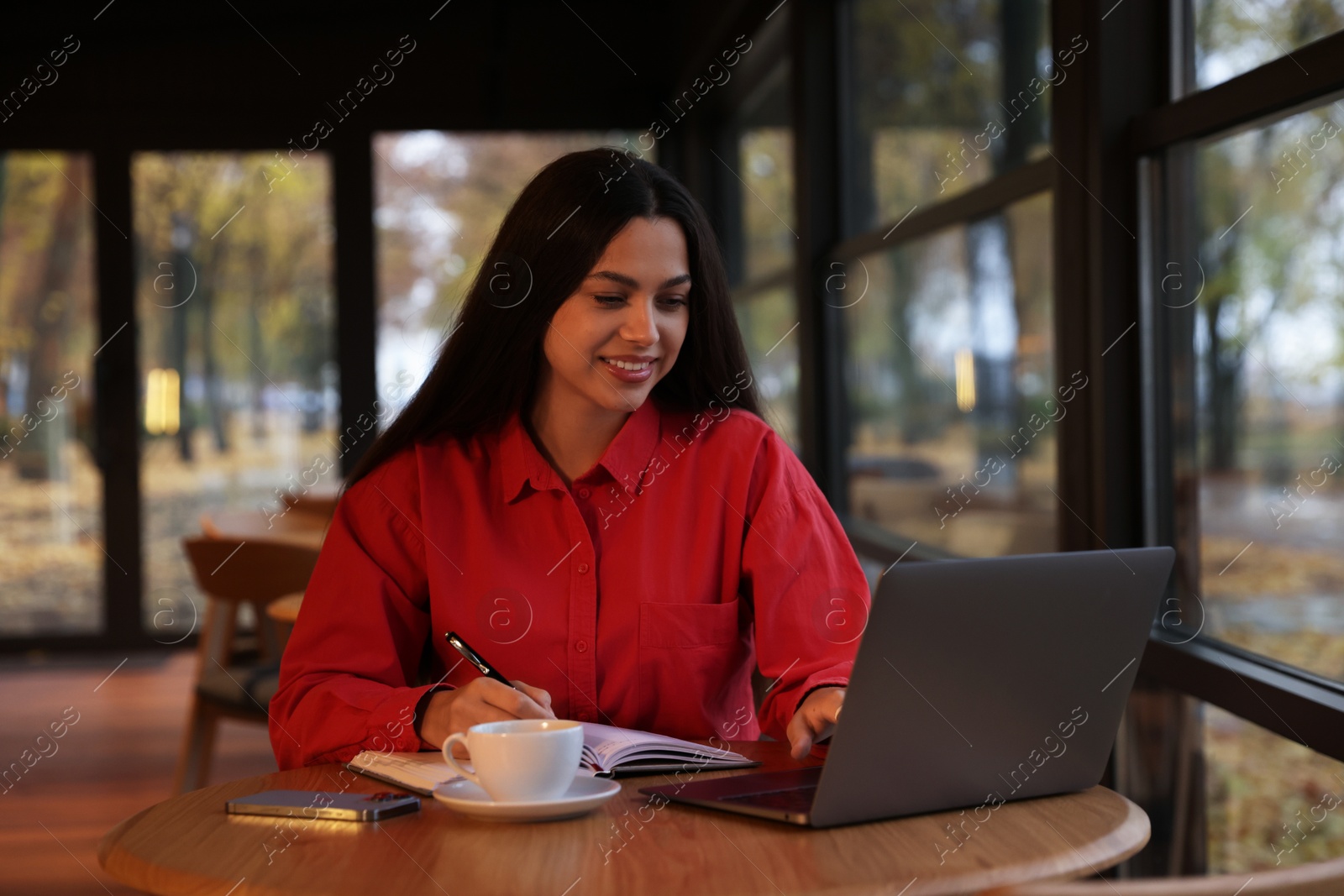 Photo of Woman taking notes while working on laptop at table in cafe