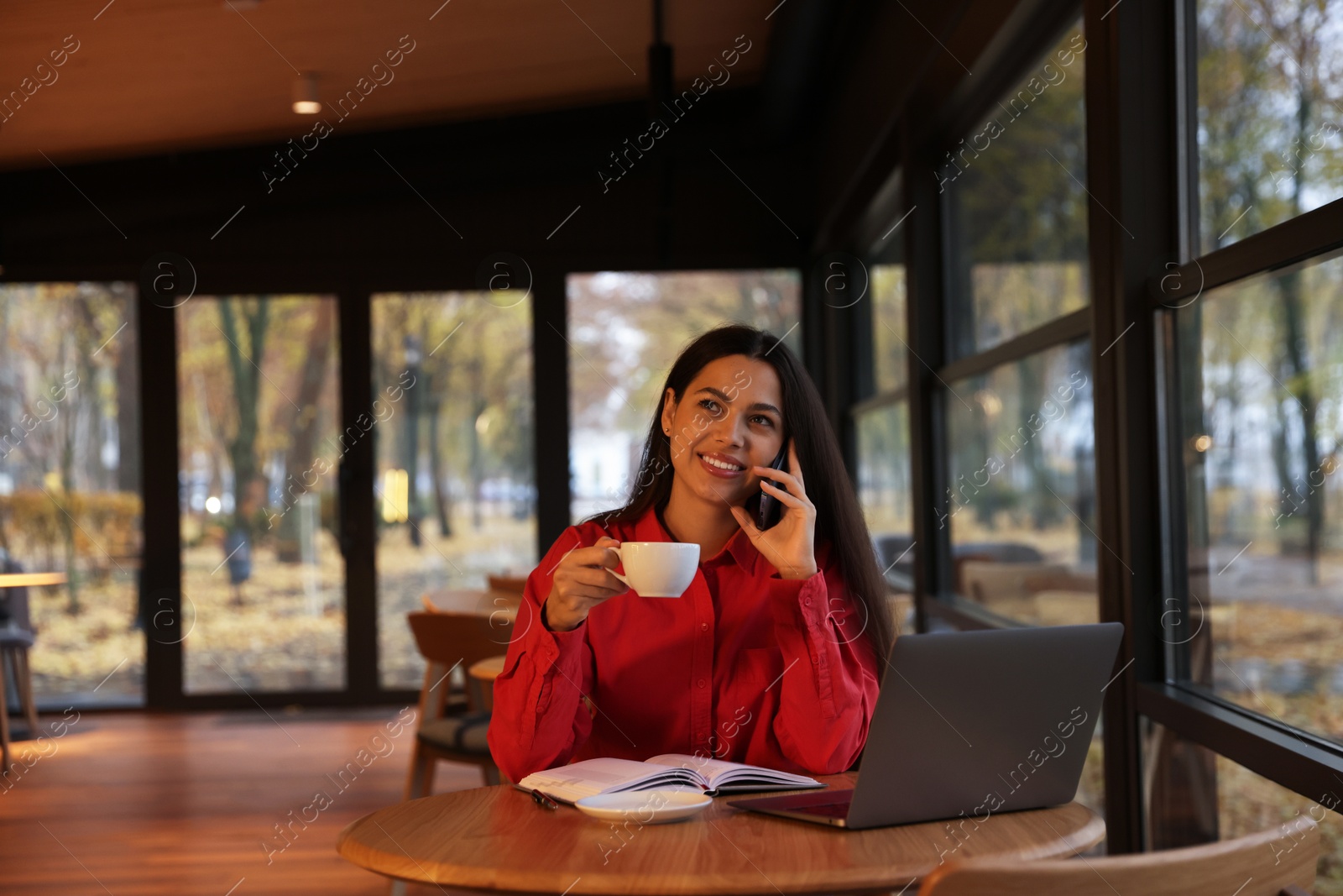 Photo of Woman with coffee talking on smartphone while working at table in cafe