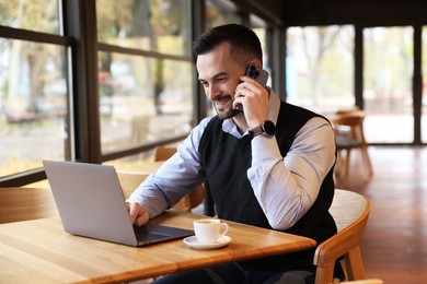 Photo of Man talking on smartphone while working with laptop at table in cafe