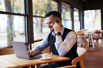 Man talking on smartphone while working with laptop at table in cafe