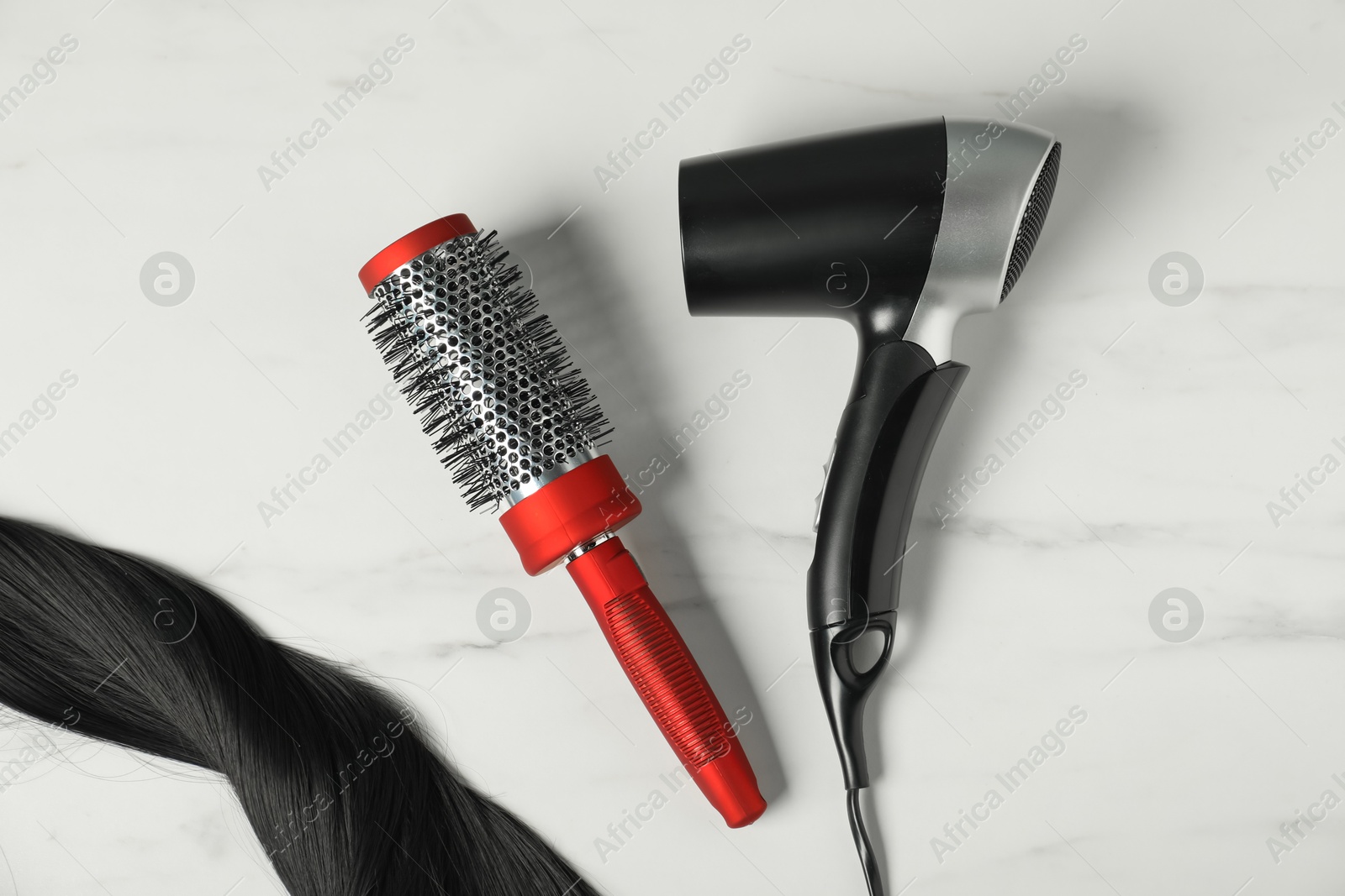 Photo of Stylish round brush, hairdryer and lock of black hair on white marble table, flat lay