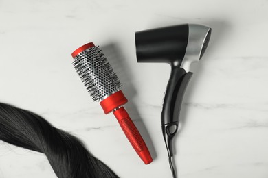 Photo of Stylish round brush, hairdryer and lock of black hair on white marble table, flat lay