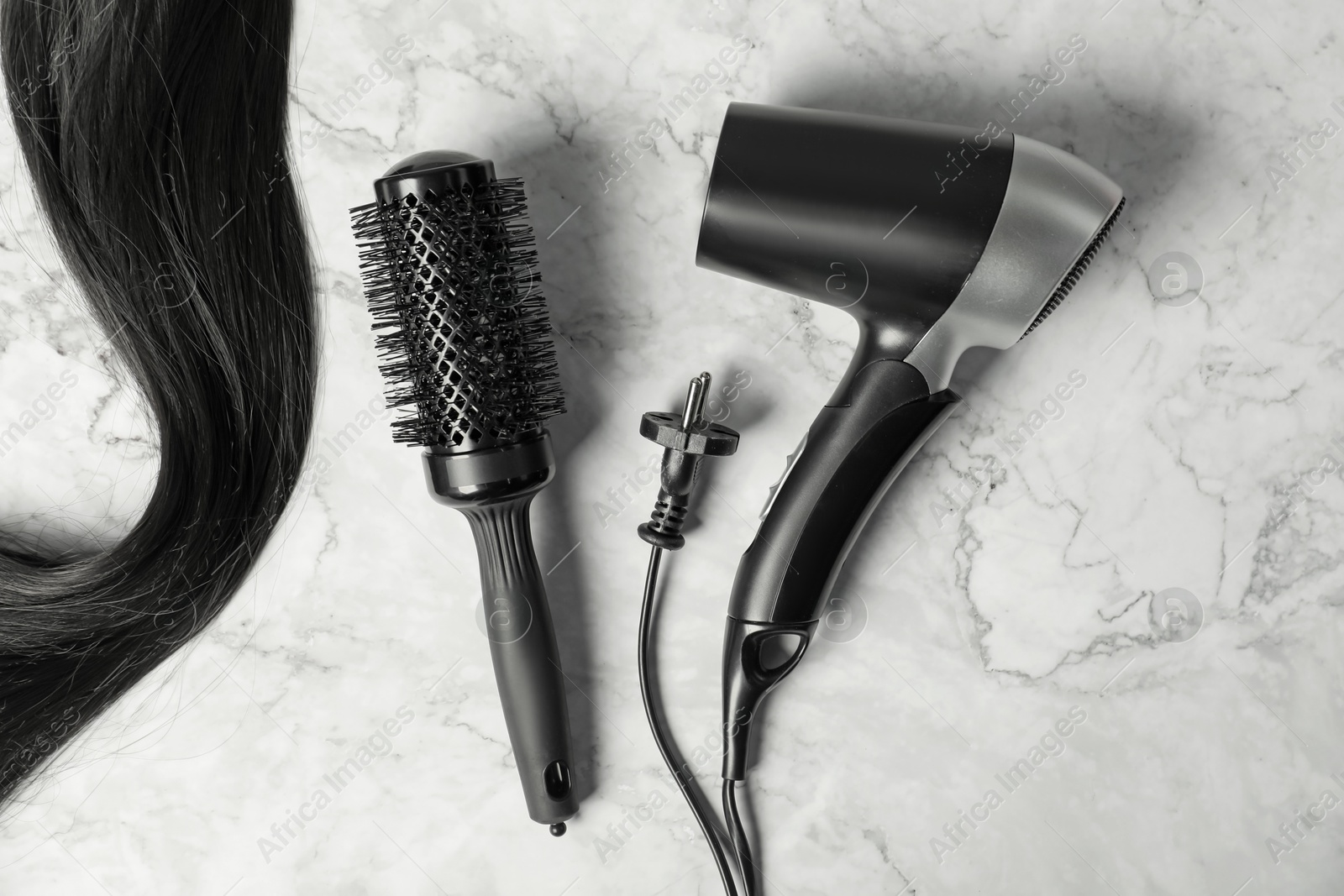 Photo of Stylish round brush, hairdryer and lock of black hair on white marble table, flat lay