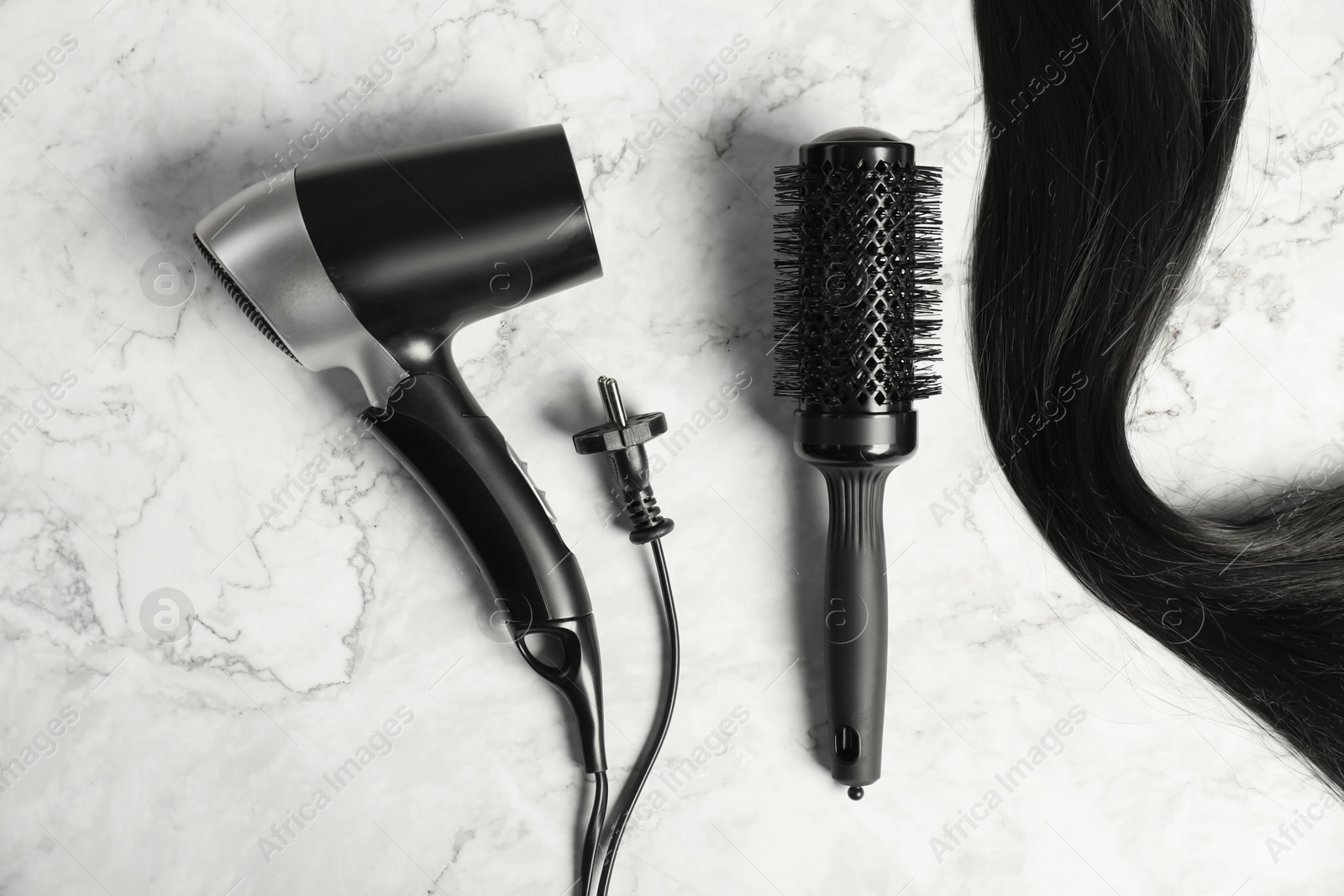 Photo of Stylish round brush, hairdryer and lock of black hair on white marble table, flat lay