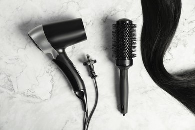 Photo of Stylish round brush, hairdryer and lock of black hair on white marble table, flat lay