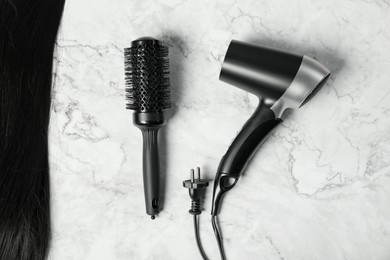 Photo of Stylish round brush, hairdryer and lock of black hair on white marble table, flat lay