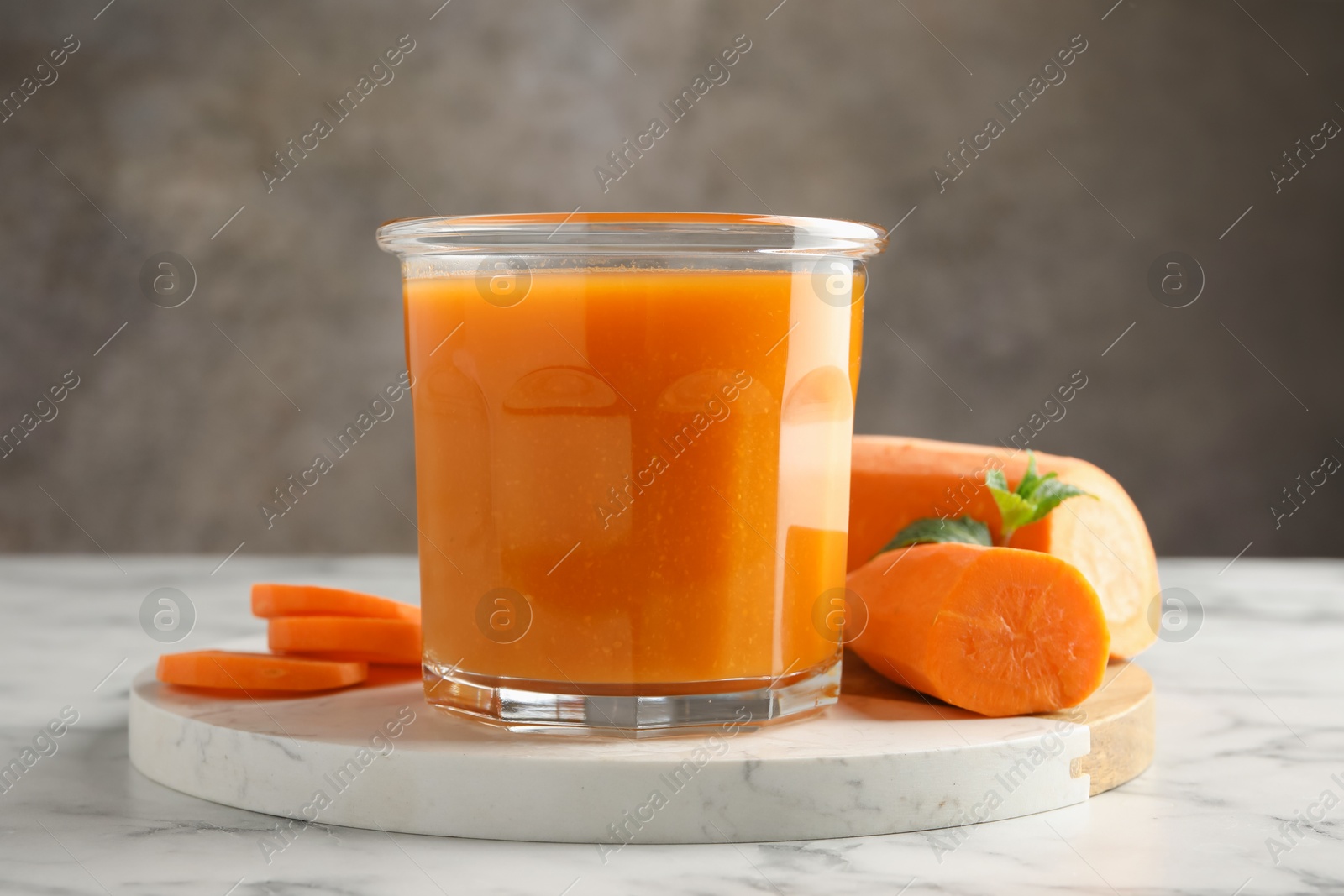 Photo of Fresh carrot juice in glass and vegetables on white marble table against gray background, closeup