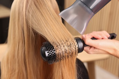 Photo of Hairdresser styling client's hair with round brush and hairdryer in salon, closeup