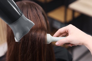 Photo of Hairdresser styling client's hair with round brush and hairdryer in salon, closeup