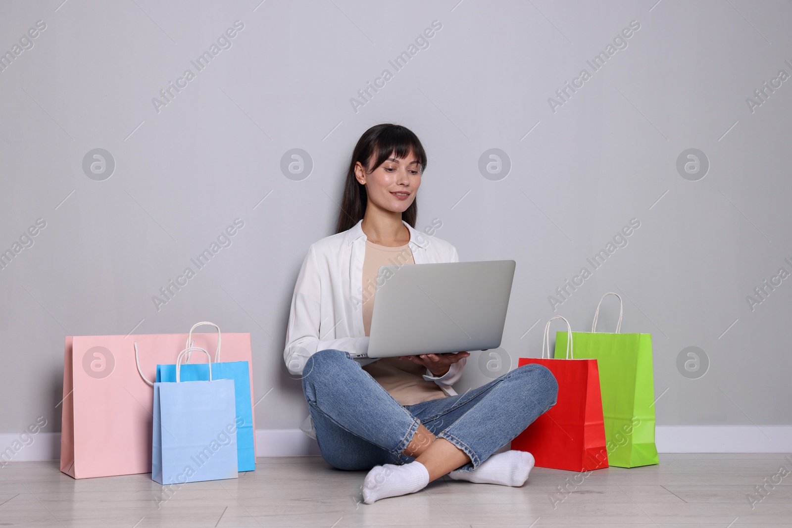 Photo of Internet shopping. Happy woman with laptop and colorful bags sitting near grey wall