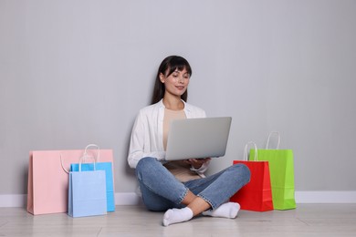 Internet shopping. Happy woman with laptop and colorful bags sitting near grey wall