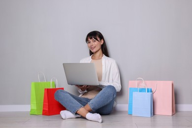 Photo of Internet shopping. Happy woman with laptop and colorful bags sitting near grey wall