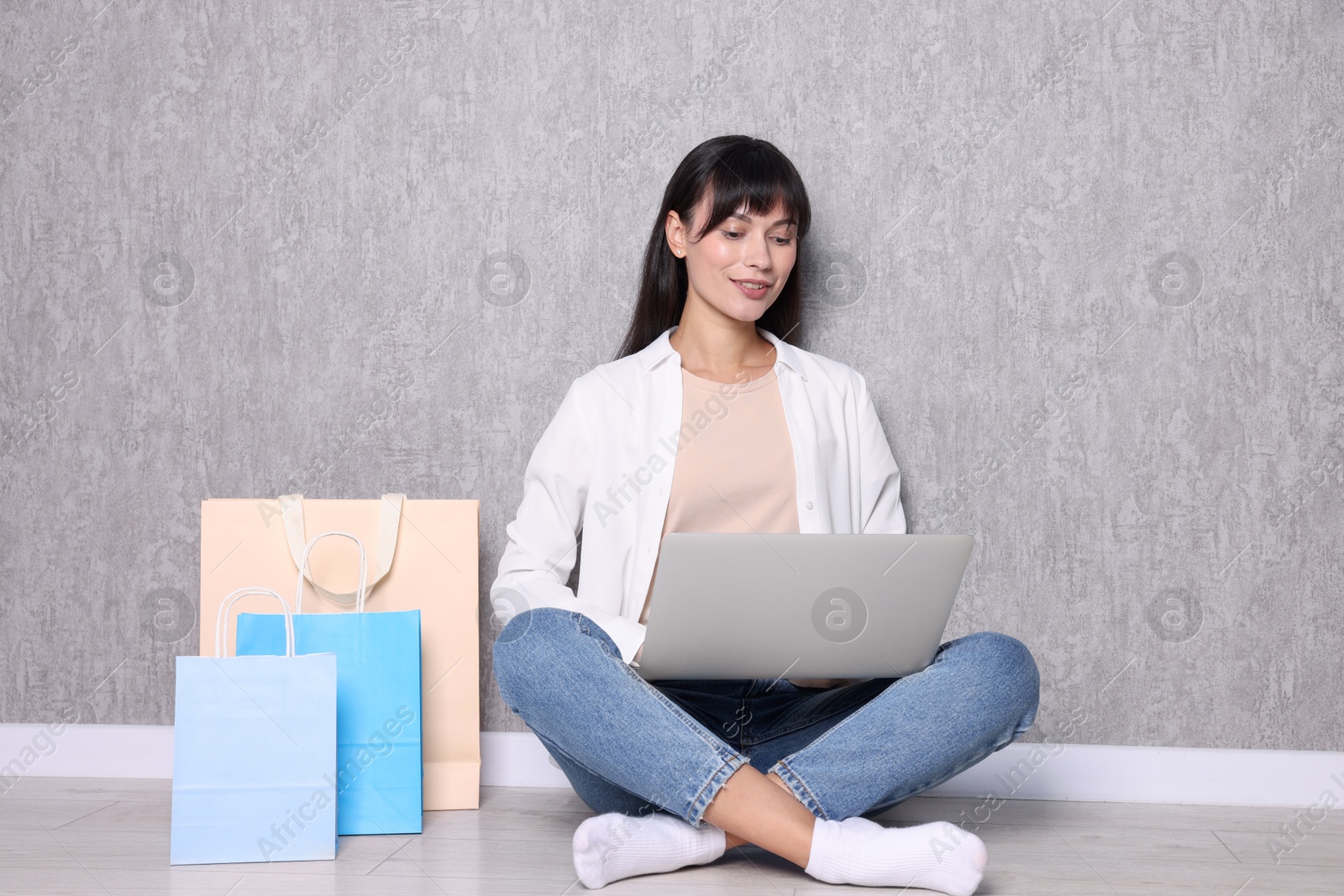 Photo of Internet shopping. Happy woman with laptop and colorful bags sitting near grey wall