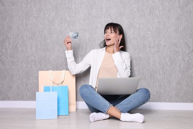 Internet shopping. Surprised woman with credit card, laptop and colorful bags sitting near grey wall